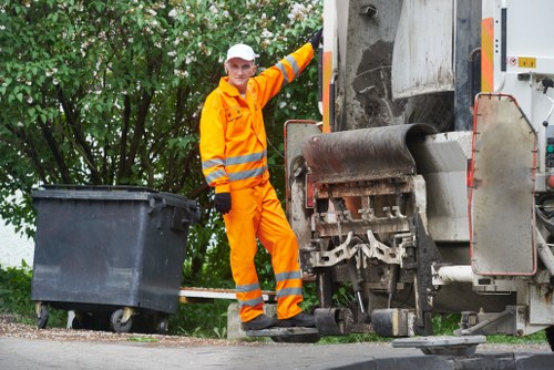 A waste collection worker dressed in a high-visibility orange uniform with reflective stripes is standing next to a large industrial rubbish collection vehicle, operated on a residential street. The worker, wearing a white safety helmet, holds onto the vehicle's side with one hand, while the other rests on a large black wheeled bin positioned near the curb. The rubbish truck, with its metallic grey body and large compactor, is parked on the roadside, partially obscured by the worker and the bin. In the background, there is greenery from trees and bushes lining the street, indicating a suburban neighbourhood. The scene is captured in daylight with natural light illuminating the area, demonstrating an organized rubbish removal operation typical in areas like Chiswick. The setting suggests tasks likely performed by local waste management services such as Garden Clearance Chiswick, supporting home or garden rubbish collection and clearance services in West London.