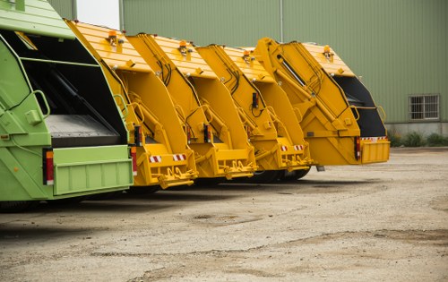 Four large industrial waste collection vehicles are parked in a line on a gravel surface in front of a green metal building with a small window. The vehicles are designed for rubbish removal, with two painted in bright yellow and one in pale green, all featuring black loading chutes at the rear for collecting waste. The yellow vehicles have reflective safety markings on their lower sections. The machinery appears clean and well-maintained, with visible hydraulic mechanisms and storage compartments. The gravel ground shows signs of frequent use, and the setting suggests this is an outdoor waste management or rubbish disposal area, possibly associated with a waste collection service in the local area, such as Chiswick. The presence of multiple vehicles indicates capacity for handling significant rubbish loads, aligning with services like those provided by Garden Clearance Chiswick for rubbish removal and waste clearance in the region.