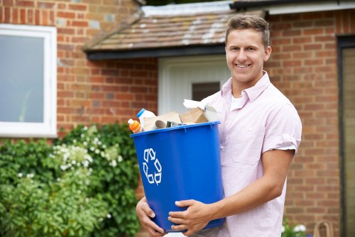 A young man with light brown hair, wearing a light pink short-sleeved shirt, is standing outdoors in front of a brick house with a white window and a small roof extension. He is holding a large blue recycling bin filled with various waste items, including paper, cardboard, and plastic bottles, which are visible at the top. The bin has a white recycling symbol on its side. Surrounding him is a garden with green foliage and white flowering plants, indicating a residential environment typical of Chiswick. The scene is illuminated by natural daylight, and the man is smiling at the camera, suggesting a positive moment of rubbish collection or garden clearance, consistent with professional waste management services offered by Garden Clearance Chiswick in the local area.