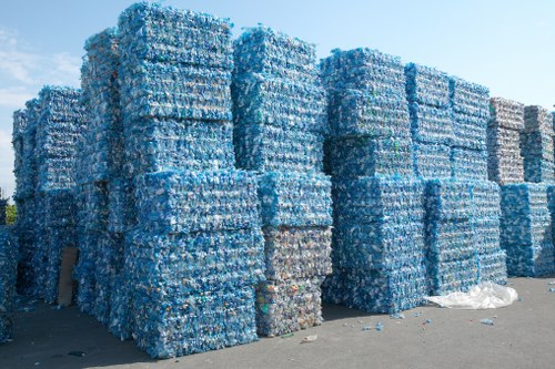 A large outdoor area showing numerous tightly-stacked pallets of crushed plastic bottles, primarily in shades of blue and transparent, arranged in multiple neat columns and rows on a paved surface. The pallets are made of wood and are stacked to several levels, creating a substantial wall of recyclable plastic waste. In the background, clear sky is visible, indicating the scene is outdoors, possibly near a recycling or waste management facility in the Chiswick area. Small amounts of plastic bottles and debris are scattered at the base of the stacks. The image emphasizes the scale of plastic waste that a rubbish removal service like Garden Clearance Chiswick might handle in a commercial or industrial context, with the focus on environmental waste management in London’s western districts.