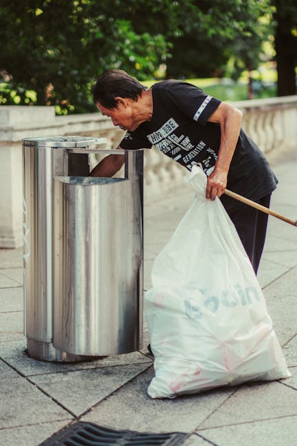 A person wearing a plaid shirt in shades of yellow, orange, and blue, along with green gloves, is holding open a large black rubbish bag made of glossy plastic material. The individual stands on a gra