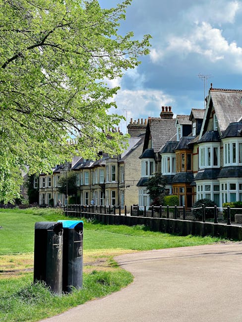 The image depicts a row of traditional terraced houses with bay windows, set on a residential street under a partly cloudy sky. In the foreground, two black waste disposal bins are positioned on a pav