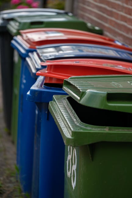 The image depicts a row of traditional terraced houses with bay windows, set on a residential street under a partly cloudy sky. In the foreground, two black waste disposal bins are positioned on a pav